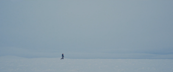 Britta Marakatt in de Finse sneeuw Beeld van witblauwe sneeuw met daarboven blauwwitte lucht, met op de achtergrond in de verte heuvels met meer sneeuw. In de verte is een kleine lopende figuur zichtbaar, in donkerblauwe warme kleding, een rode muts en witte wanten.